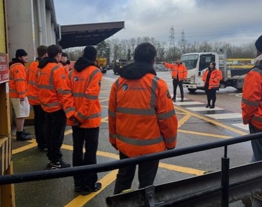 A group of young learners stand outside a depot in orange high vis jackets. 